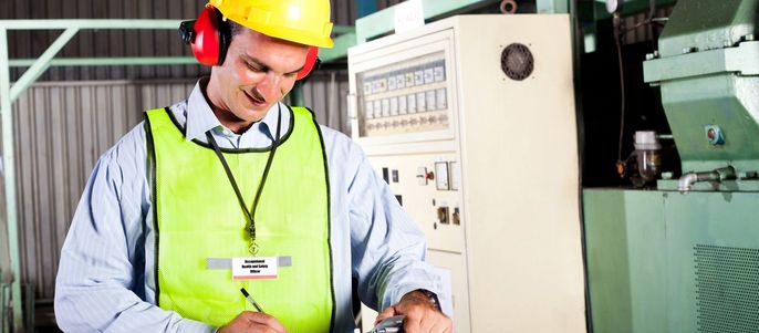 Ein Mann, der einen gelben Schutzhelm und einen Gehörschutz trägt, arbeitet in einer industriellen Umgebung, mit einer Maschine im Hintergrund.