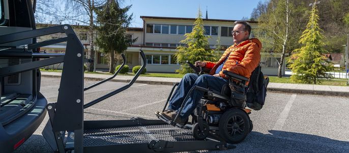 Ein Mann in einer orangefarbenen Jacke, der in einem Rollstuhl sitzt und von einer Vorrichtung unterstützt wird, um aus einem Fahrzeug zu steigen.