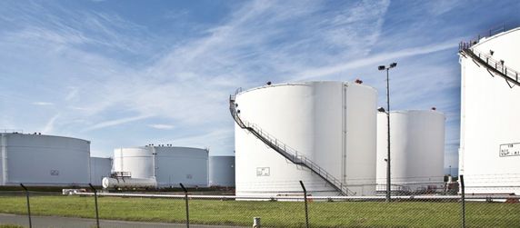 Mehrere große weiße zylindrische Lagertanks, einige mit Leitern, auf einer offenen Fläche mit einem Zaun im Vordergrund.